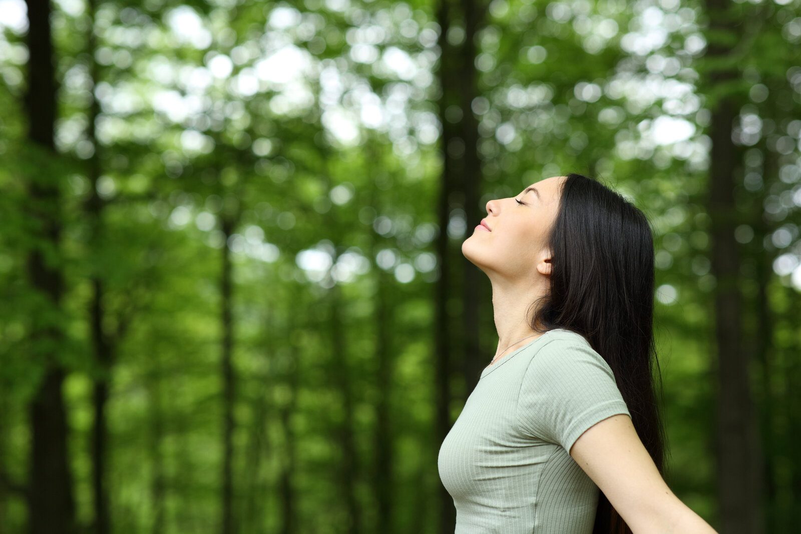 Woman standing among green forest trees with her face tilted upward